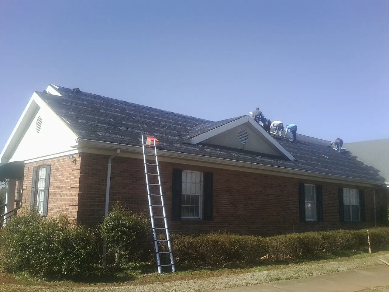 Crew tearing shingles off a brick ranch roof with a ladder set — Emergency Roof Repair work by our crew in Altamahaw