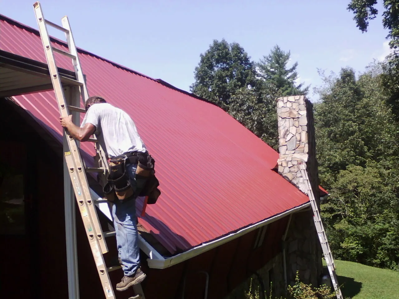 Roofer on a ladder beside a red metal roof and stone chimney — Soffit & Fascia Repair by Roofing Brothers in Altamahaw